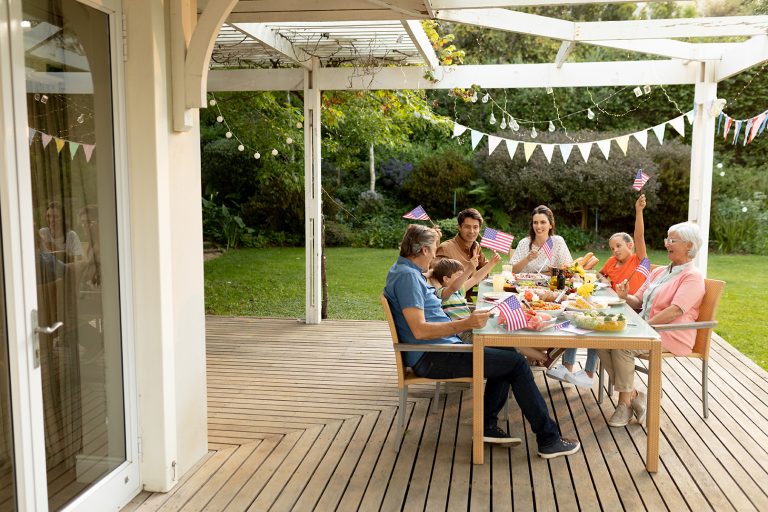 front view of a multi generation caucasian family sitting outside on a deck at a dinner table set for a meal, celebrating and waving us flags. family enjoying time at home, lifestyle concept